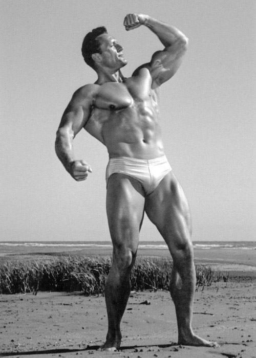 Clarence Ross on the beach posing flexing with bicep looking towards the sky in swimming shorts