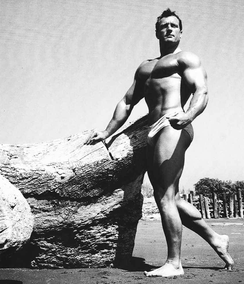 Clarence Ross standing next to a rock on the beach in swimming shorts with strong muscles 