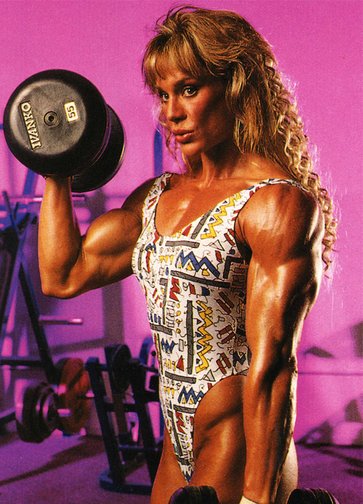 Corinna Everson lifting weights in the gym, displaying her muscular arms.