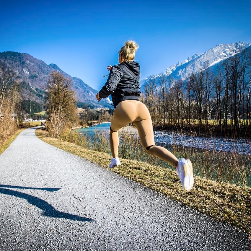 Alexandra Bring in light-brown leggings, running outside by a river stream, in a picturesque landscape with mountains and withered trees in the background