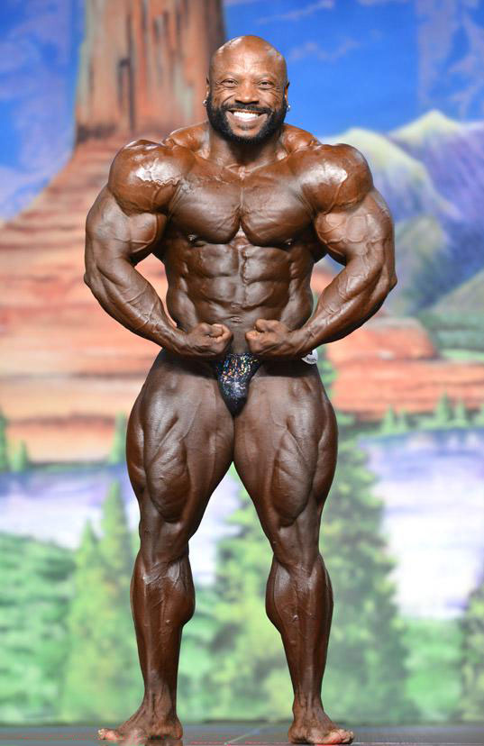 Charles Dixon standing on a bodybuilding stage flexing his huge muscles while smiling