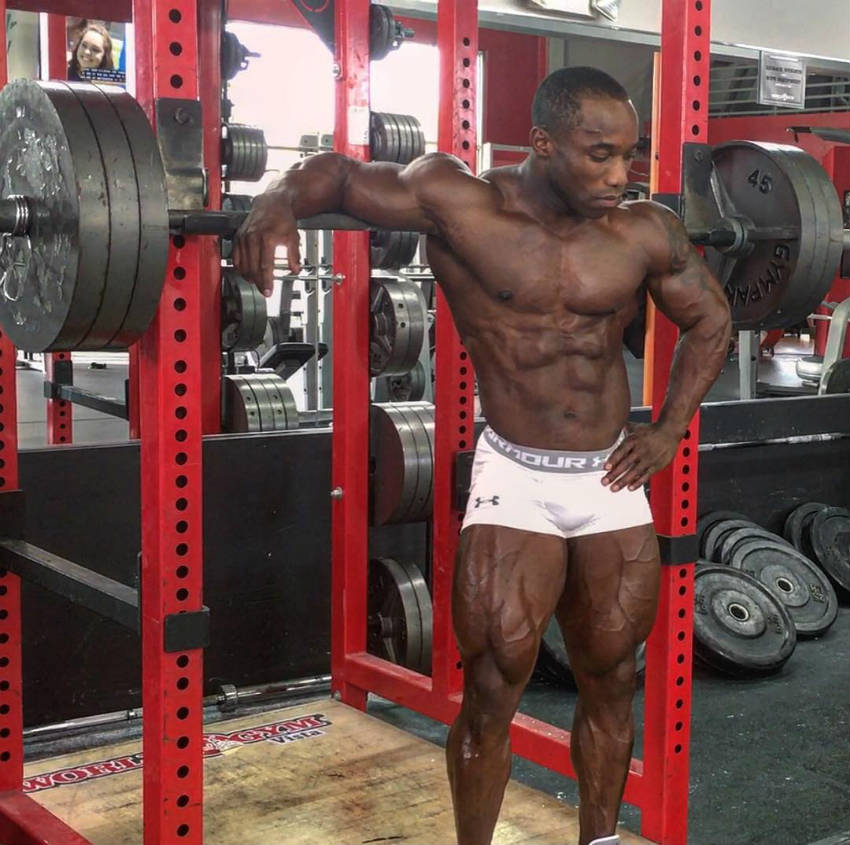 Damion Rickett standing in front of a barbell on the rack, showing his large quadriceps and ripped abs