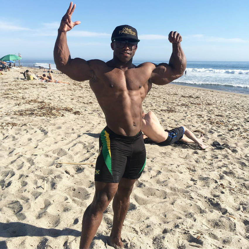 Damion Ricketts standing on the beach showing his full body and tensing his biceps