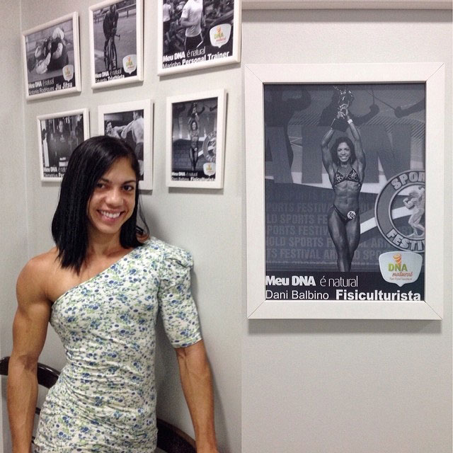 Dani Balbino wearing a white dress standing next to her own picture n the wall highlighting her bodybuilding competition achievements