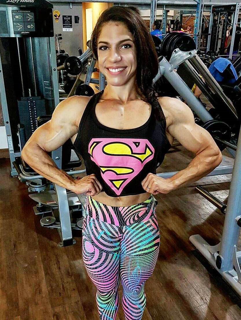 Dani Balbino standing in the gym flexing both of her biceps and chest