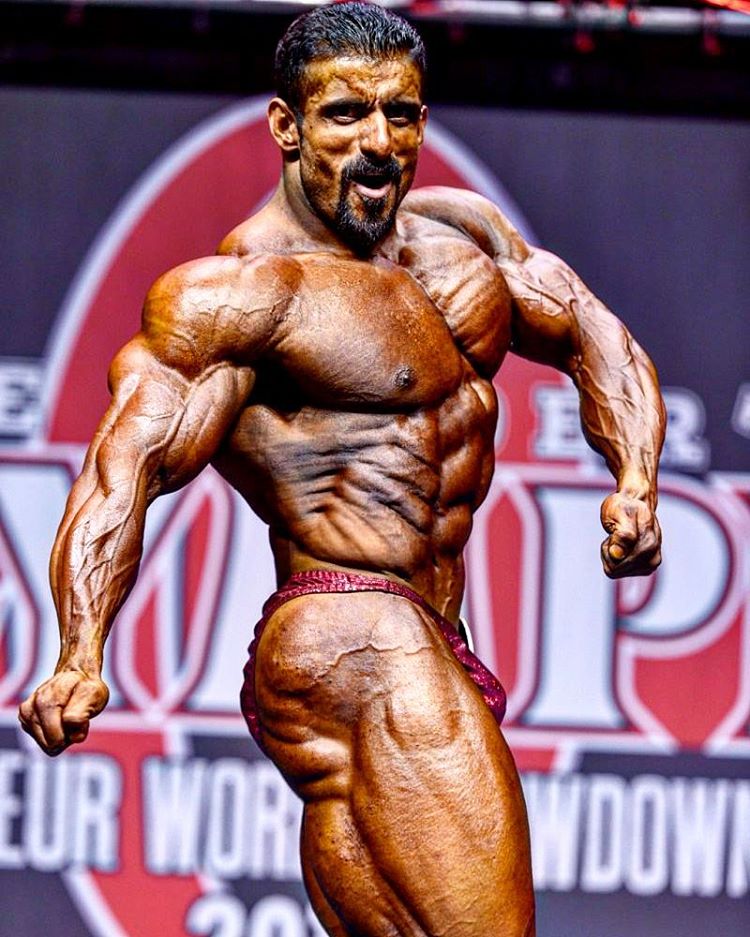 Hadi Chopan on the Olympia stage, side pose, flexing his arms, chest, obliques, legs, and glutes, displaying his incredible conditioning