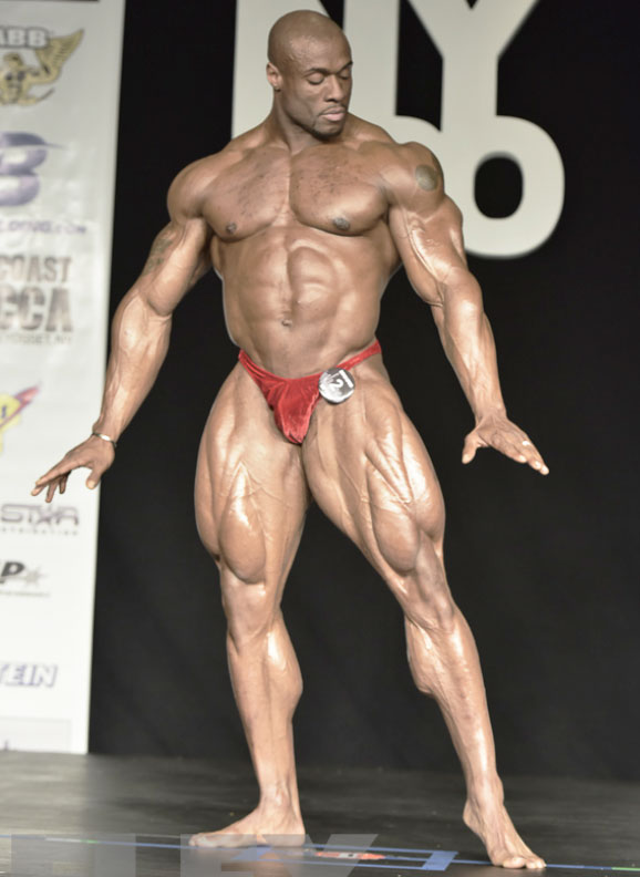 Jeff Beckham posing on a bodybuilding stage in red trunks pointing towards the ground with both hands 