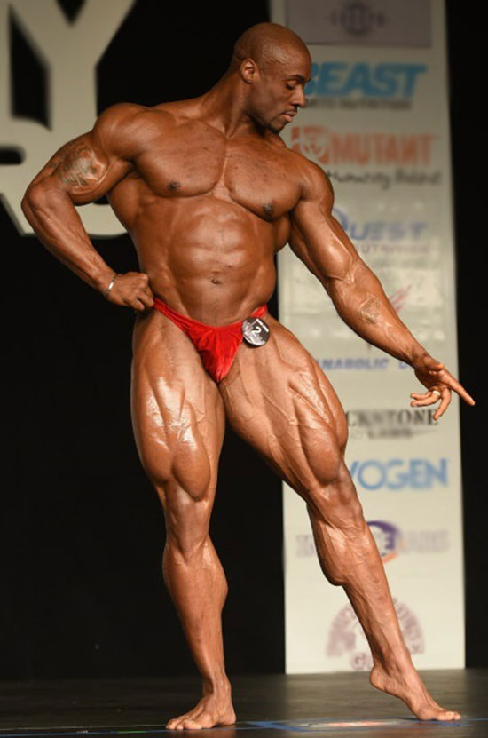 Jeff Beckham posing on a bodybuilding stage in red trunks pointing towards the ground 