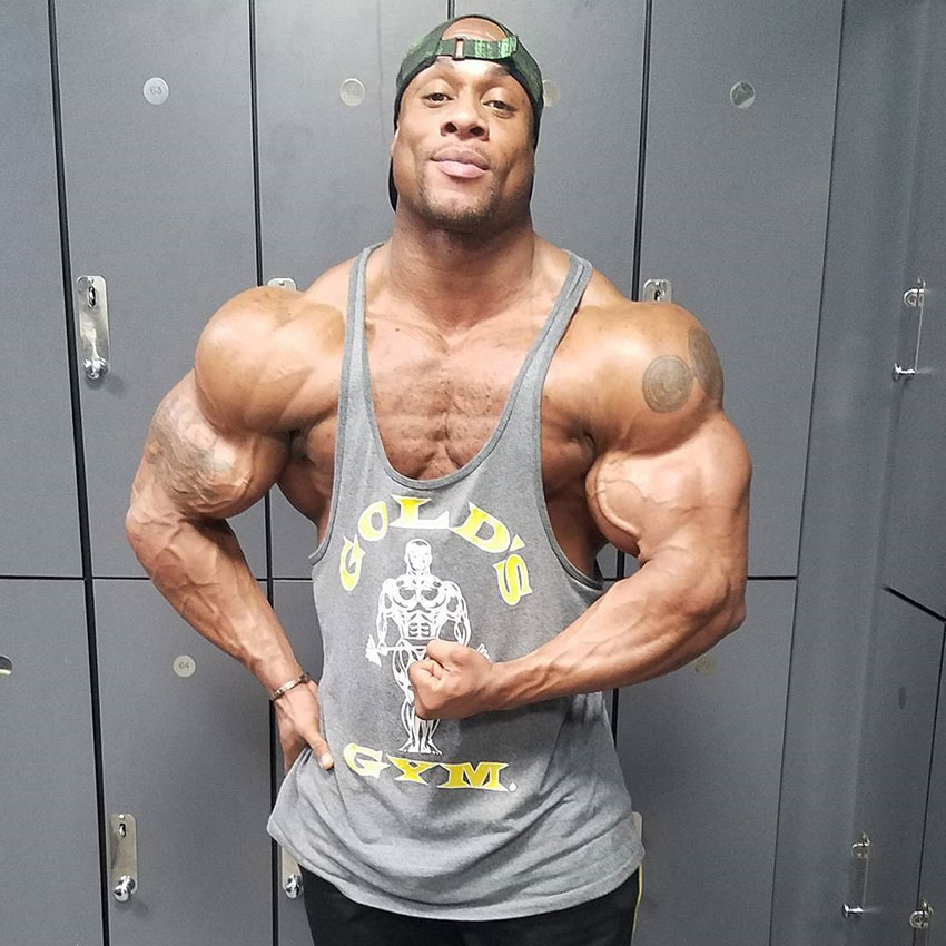 Jeff Beckham standing in the gym changing room in front of lockers flexing his left bicep looking huge and strong 