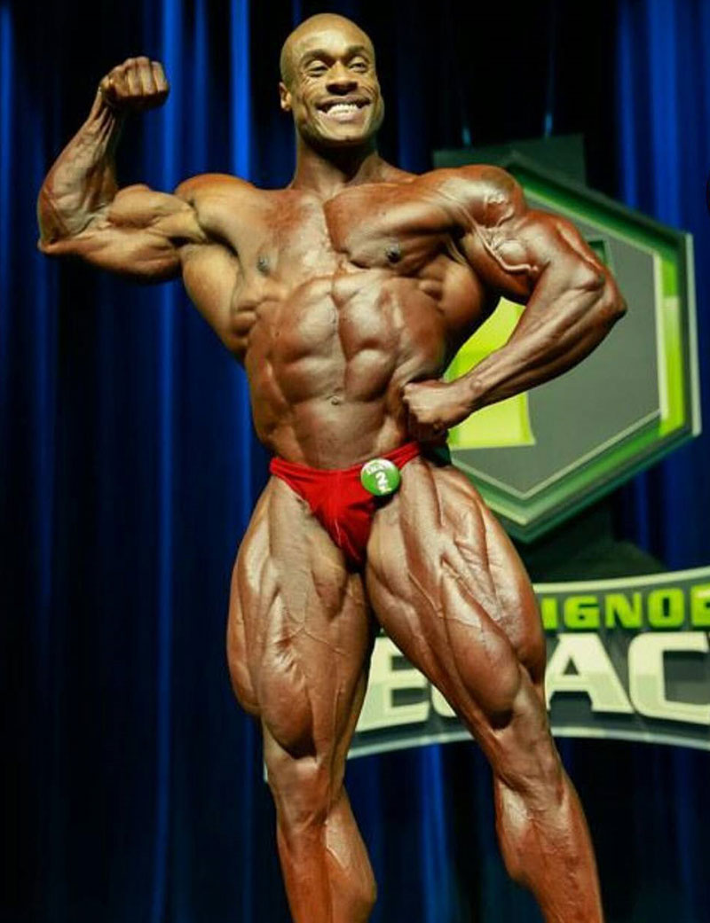 Jeff Beckham posing on a bodybuilding stage flexing his right bicep smiling wearing red trunks 