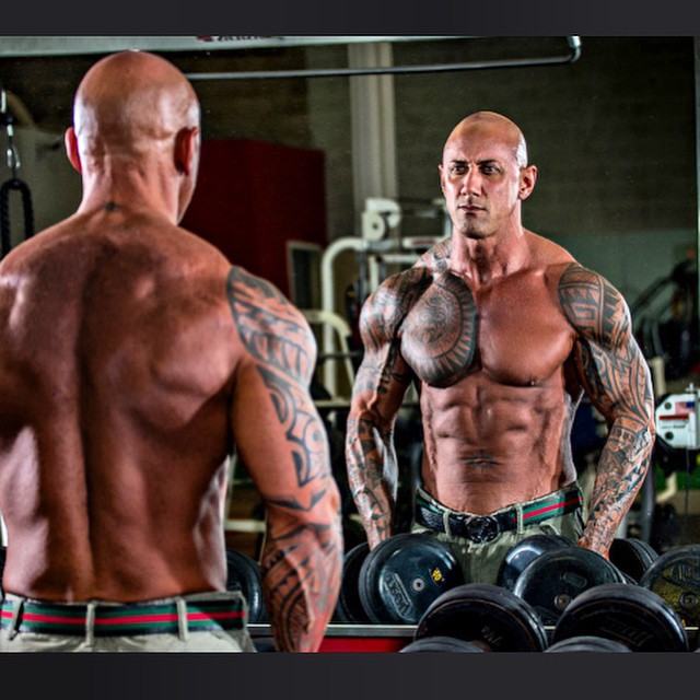 Shirtless Jerry Ward looking at himself in the gym mirror while holding dumbbells in his hands