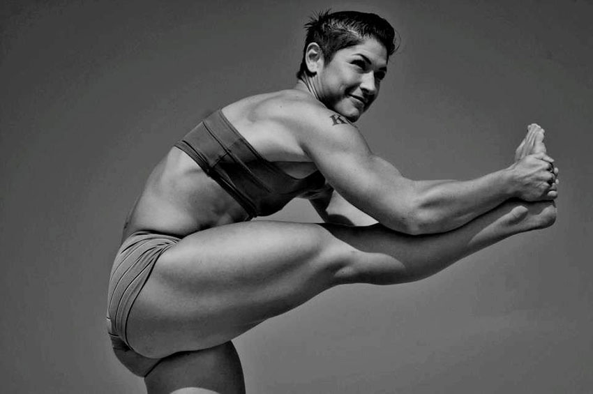 Kortney Olson standing on one leg while stretching her other leg showing her large leg muscles and muscular body 