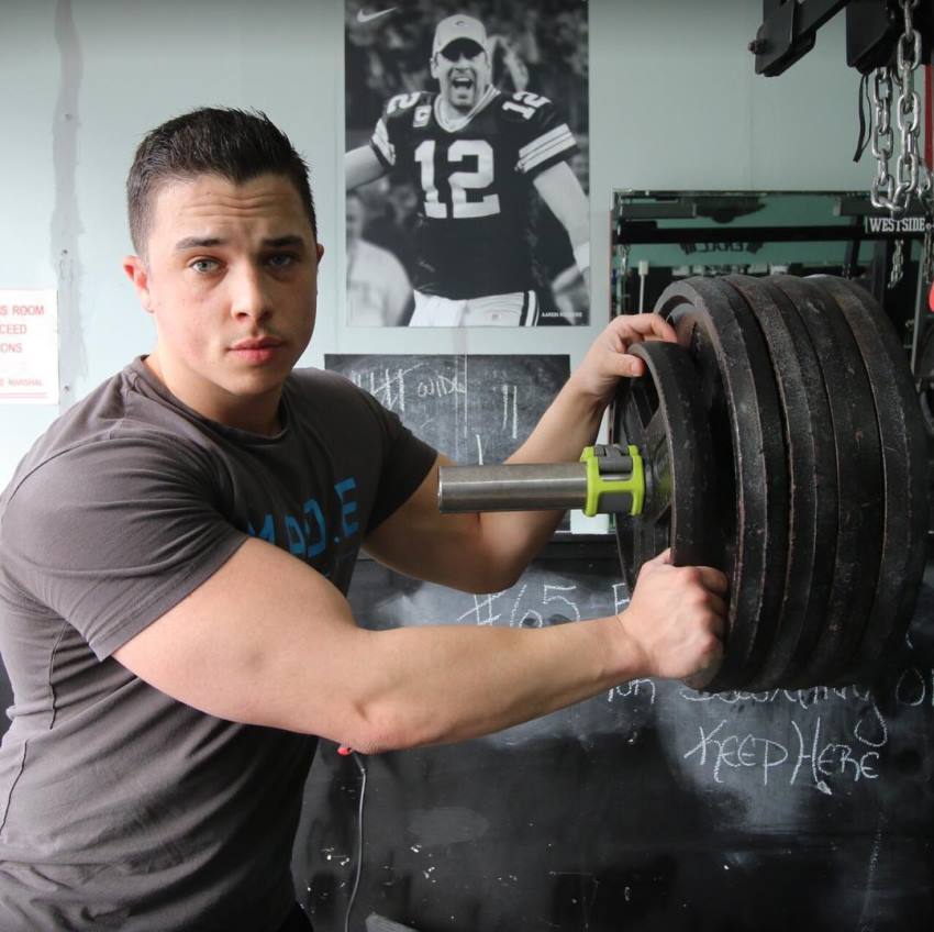 Nick Whright putting a lot of weight on a weight bar, preparing to do a heavy lift, as she looks straight into the camera, and flexes his arms