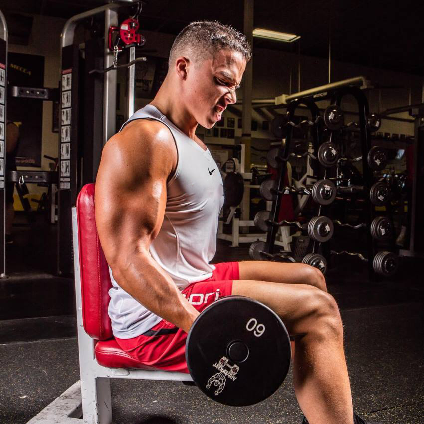 Side show of Nick Wright performing seated dumbbell biceps curls in a white tank top