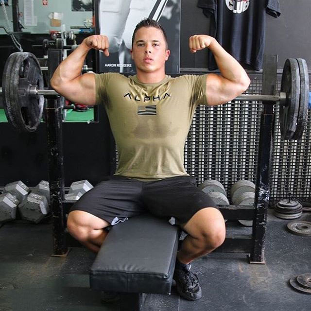 Nick Wright sitting on a bench in the gym flexing both biceps 