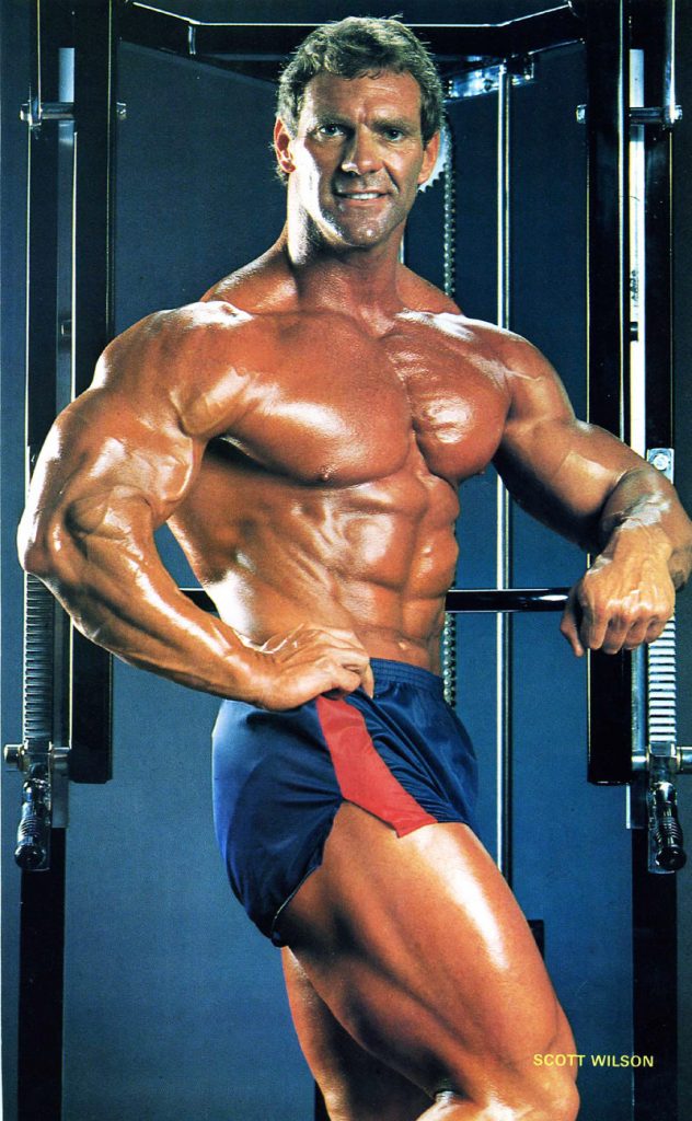 Scott Wilson posing in blue and red shorts, tensing his abs and upper body, while displaying his muscular legs.