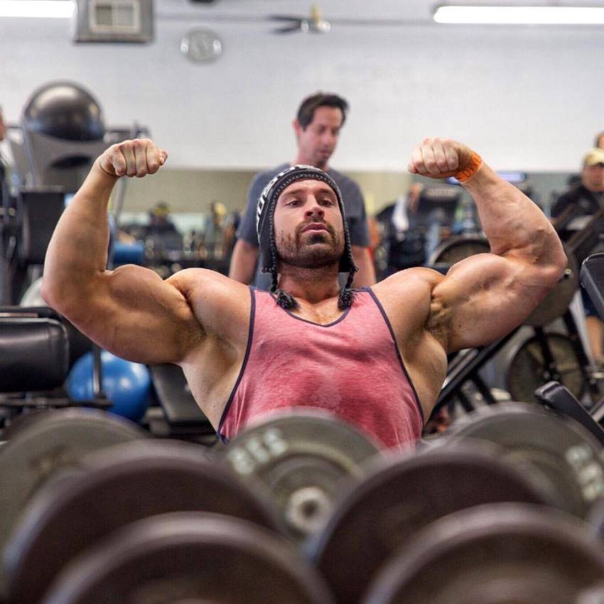 Bradley Martyn in einer vorderen doppelten Bizeps-Pose, während er auf einer Bank in einem Fitnessstudio sitzt und ein rotes Tank-Top trägt