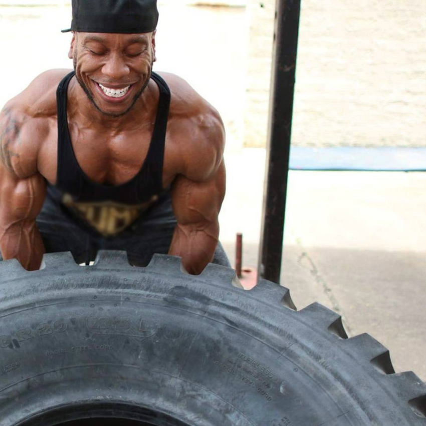 Chris Jones flipping a big tire while being in black and gold tank top, showing his incredibly ripped arms, shoulders, and pecs