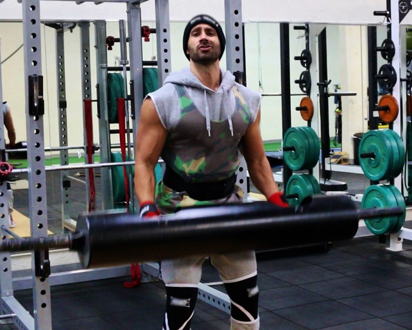 Dom Mazzetti lifting a black weight bar while saying something sarcastic into the camera