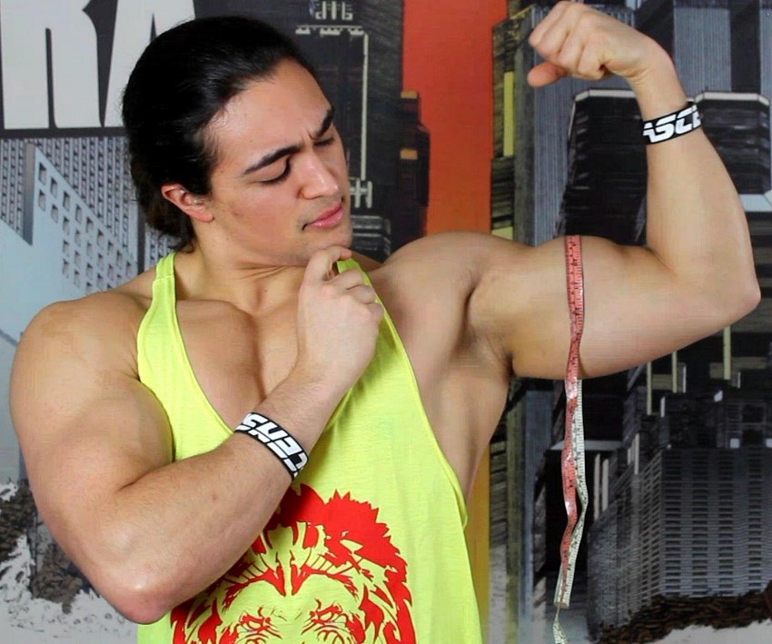 Omar Isuf in a yellow tank top, measuring his flexed biceps