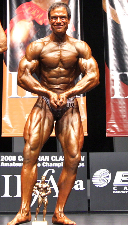 Rob Deluca on the stage with a trophy on the floor, showing his contest-winning physique in a most muscular pose