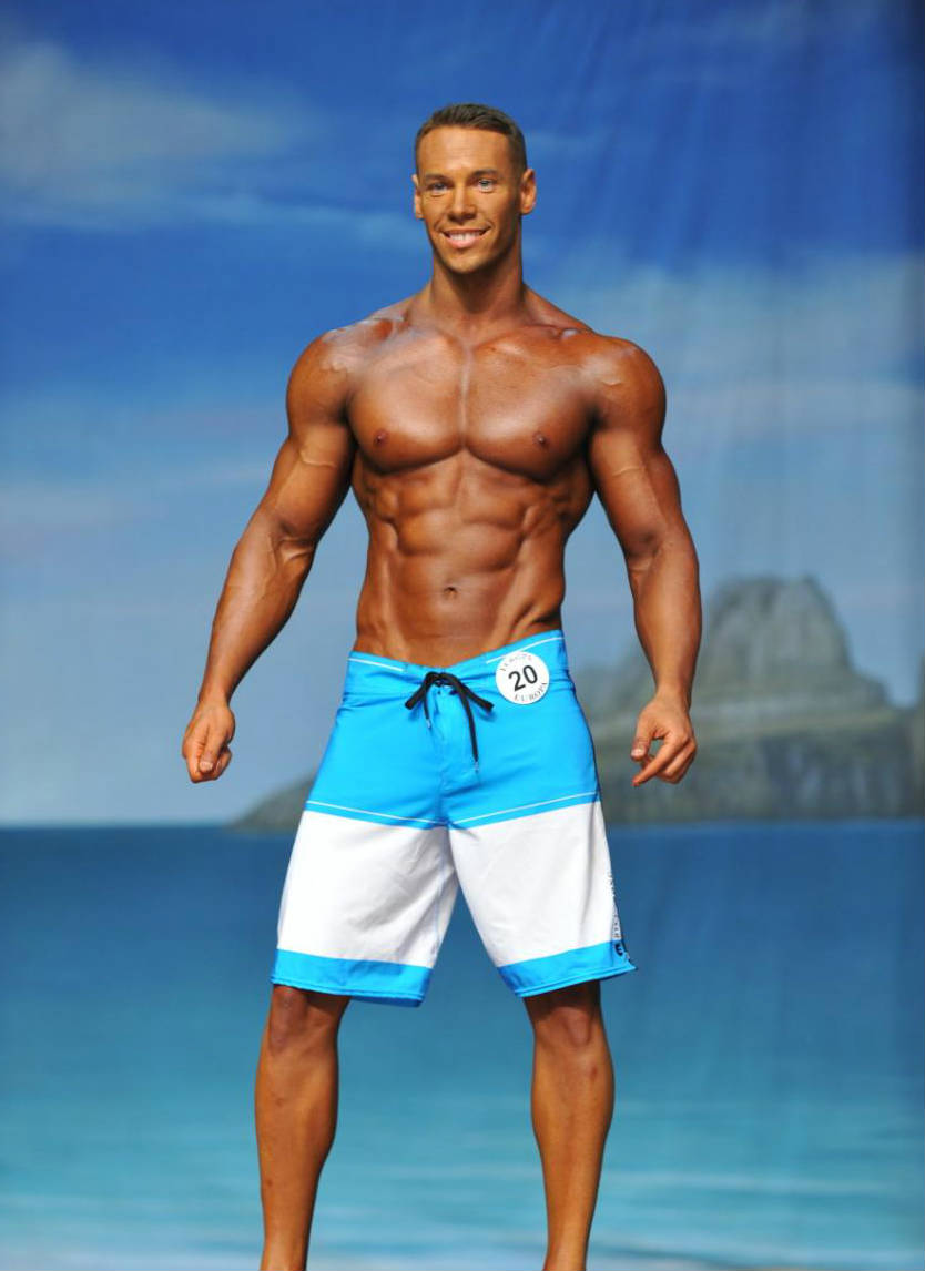 Anton Antipov standing n board shorts at a competition, showign off his reipepd abs, toned obliques and large chest and delts