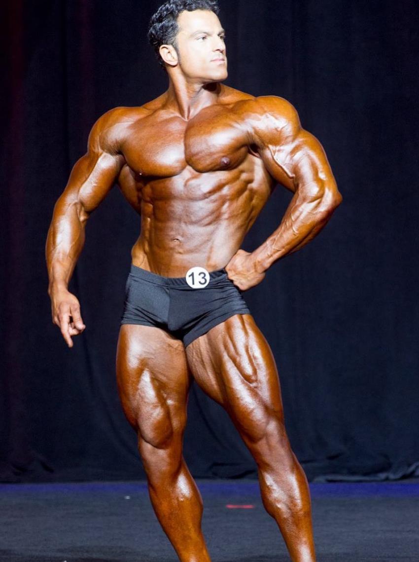 Arash Rahbar confidently posing on the stage in black trunks, showing his classic golden era physique to the judges