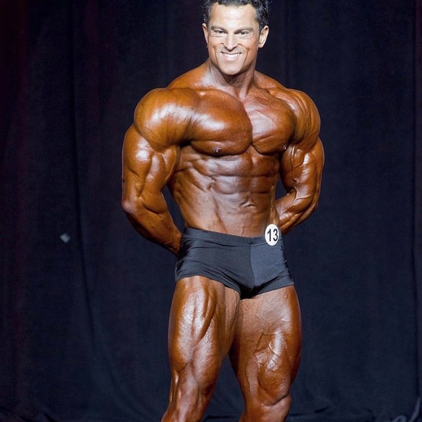 Arash Rahbar showing a variation of his most muscular pose to the judges on the stage, smiling at them, and being confident in his impressive physique