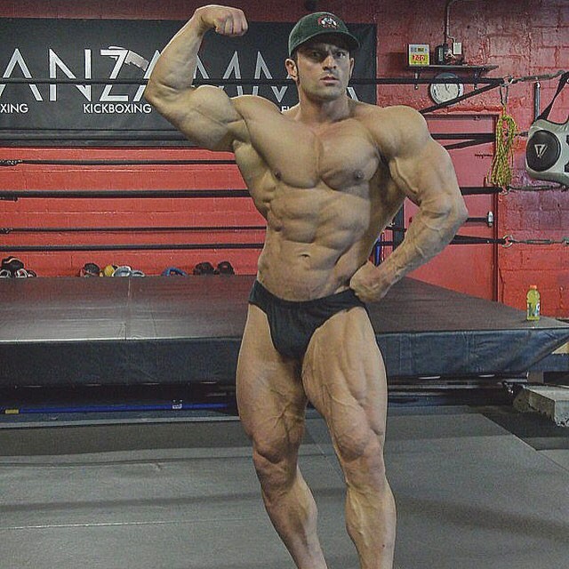 Arash Rahbar standing in front of a boxing ring in his black trunks, flexing his biceps, and showing his shirtless ripped body