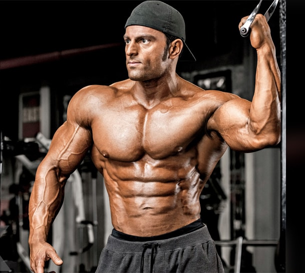 Arash Rahbar doing a photoshoot in the gym, holding onto a cable, and showing his ripped and muscular body