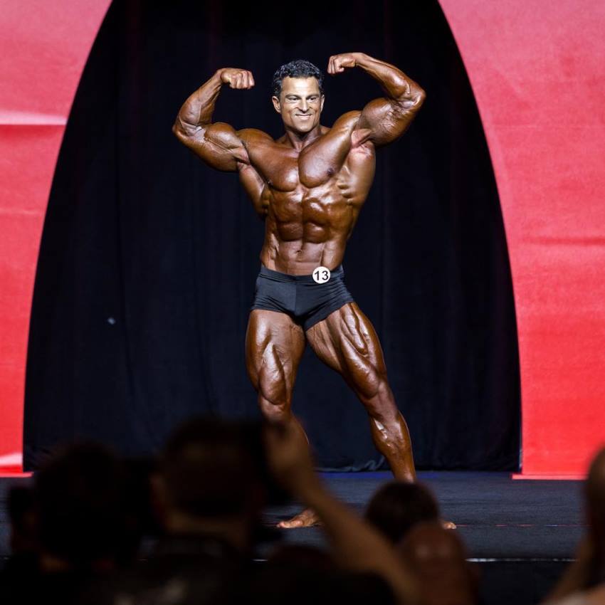 Arash Rahbar doing a double biceps flex pose on the olympia stage, smiling at the photographers, judges, and fans who are cheering him up in the background