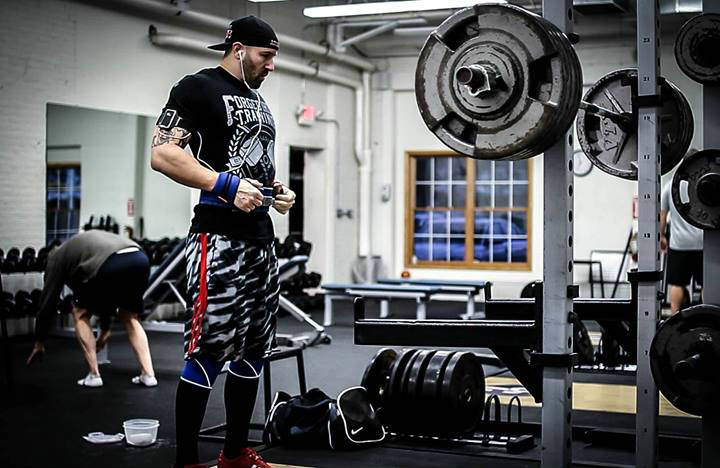 Brandon Campbell standing in the powerlifting gym, showing his large arms and legs 