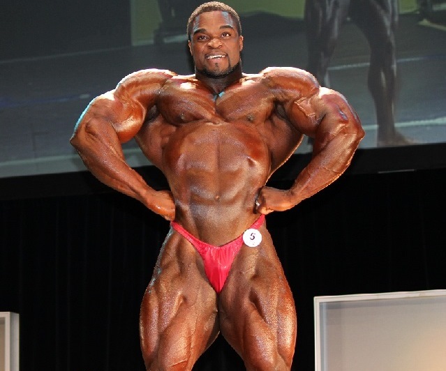 Brandon Curry in the front lat spread pose in red bodybuilding underwear, showing is big and muscular physique