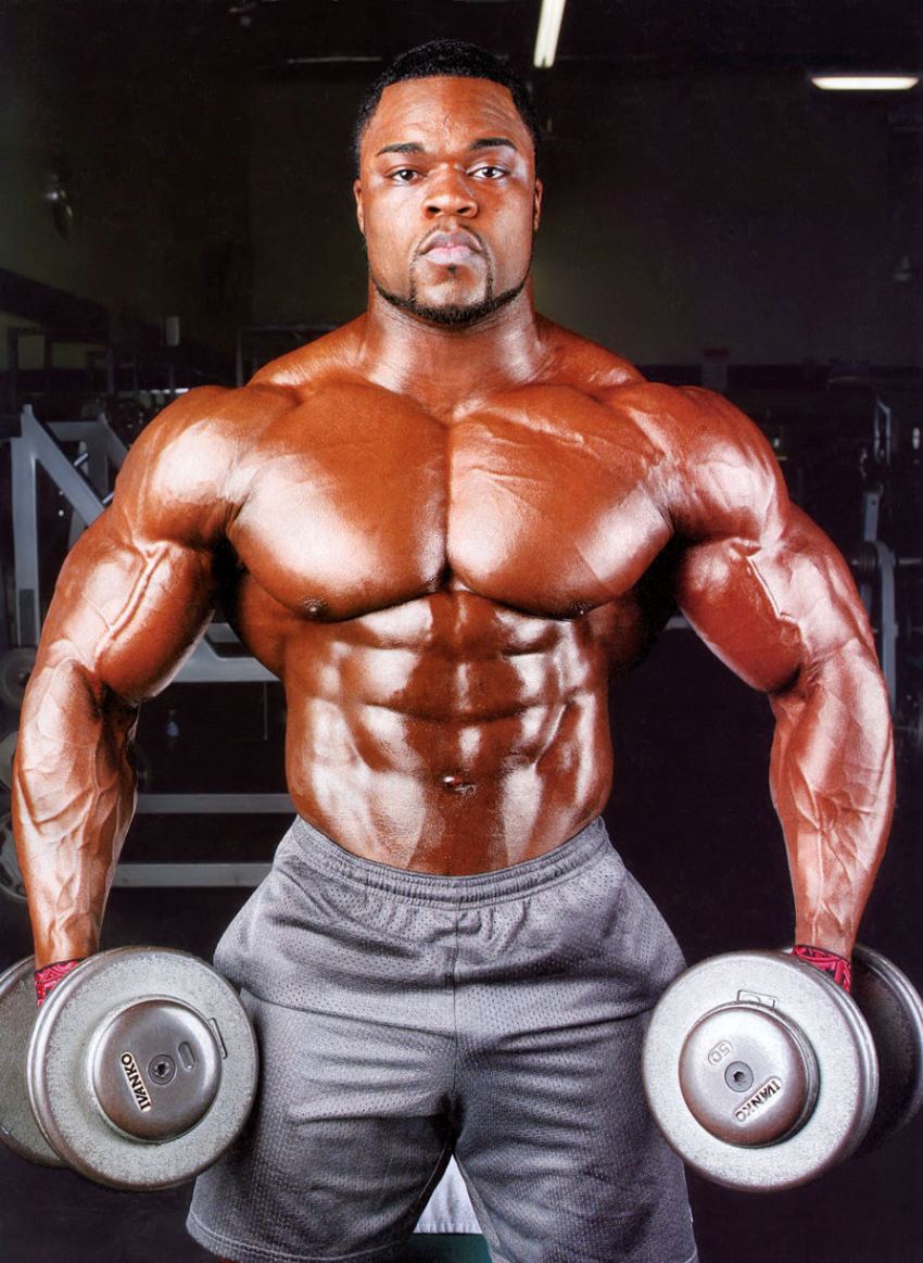 Brandon Curry shirtless in grey shorts, doing side lateral raises for his strong and big shoulders