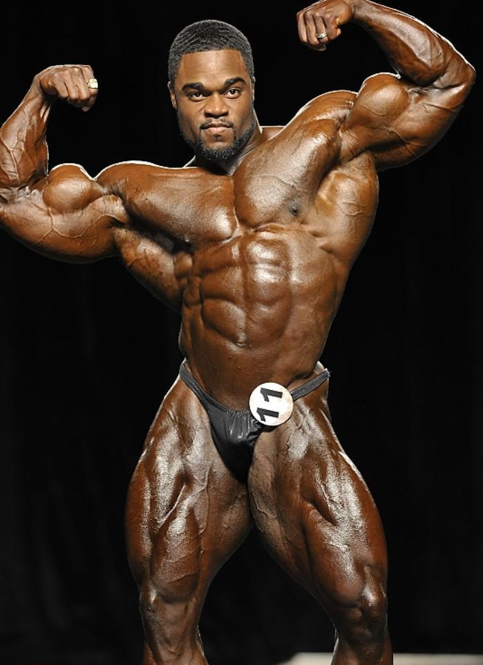 Brandon Curry on the stage, in a front double biceps pose, displaying his impressive conditioning to the judges