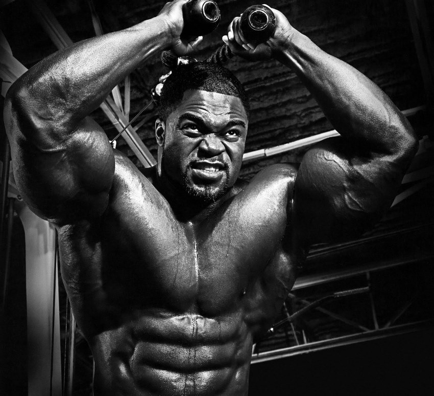 Brandon Curry doing overhead cable extensions for his triceps, as he flexes his ripped abs, chest, and arms