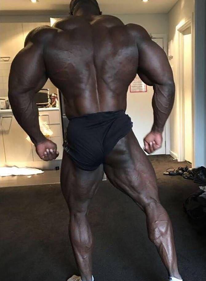 Brandon Curry standing in black shorts, showing his incredible back, arms, and legs development