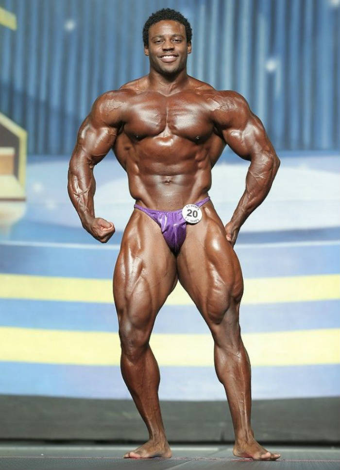 Breon Ansley standing with a confident grin at a competition showing his well-built quads