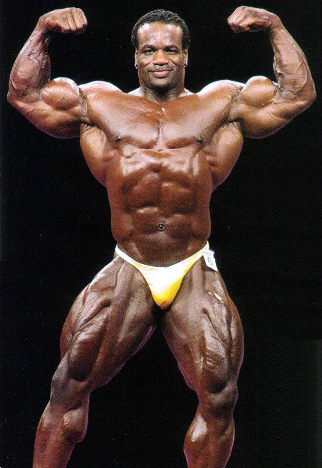 Chris Cormier posing at a competition, showing his large quads, ripped abs and large bulging biceps