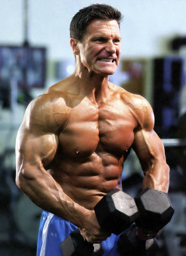 Clark Bartram listing two dumbbells and showing his large arms, chest and abs 