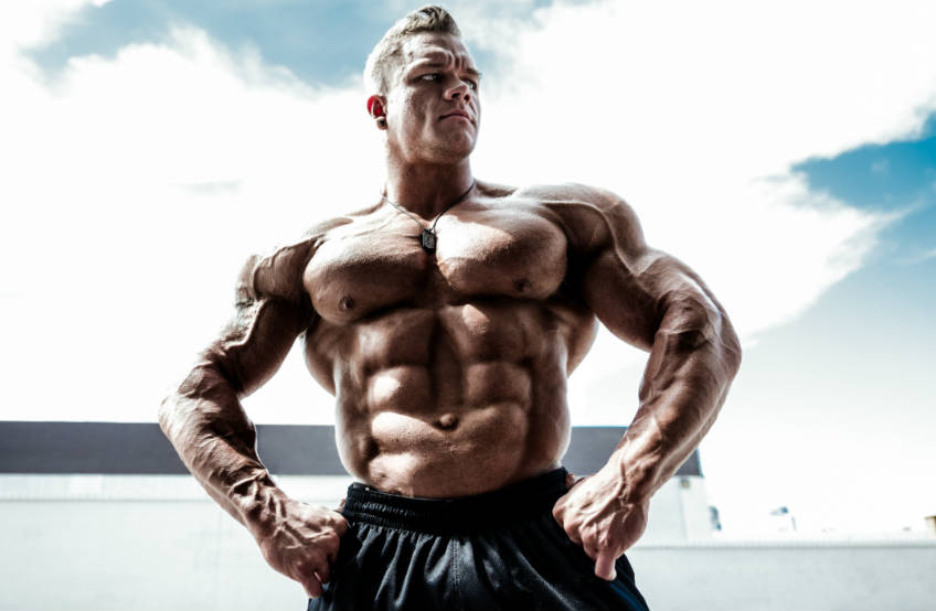 Dallas Mccarver posing with his hands on his hops, showing off his ripped abs and massive chest