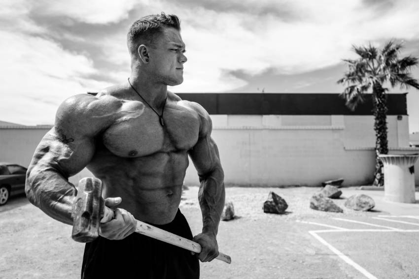 Dallas Mccarver standing with a sledge hammer, showing his large chest, arms and ripped abs 