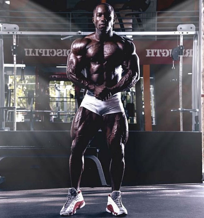 Damion Ricketts flexing his muscles in the gym wearing gym white gym shorts looking lean and muscular