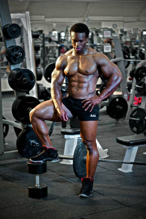 Daron Lytle standing on a dumbbell in the gym, showing his ripepd abs, large arms and chest and toned legs