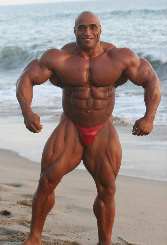 Dennis James standing on the beach, showing his large chest, arms and quads