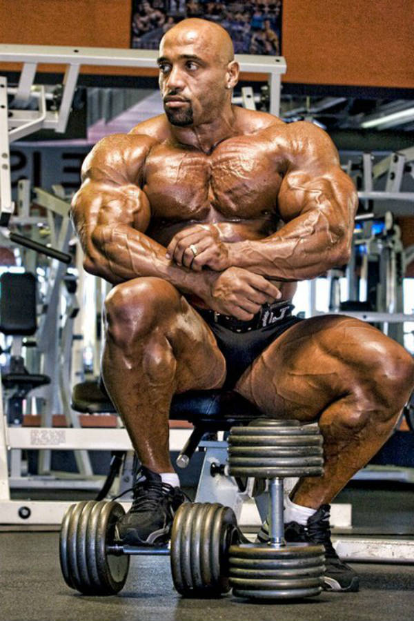 Dennis James sitting by dumbbells, showing his vascular physique and large arms