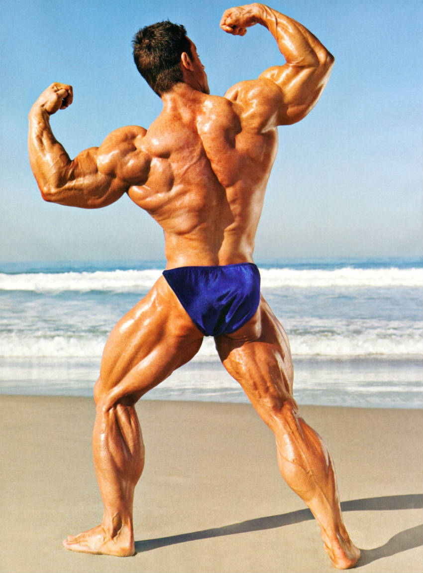 Erik Fankhouser standing on the beach showing his back profile to the camera and flexing his arms and back 