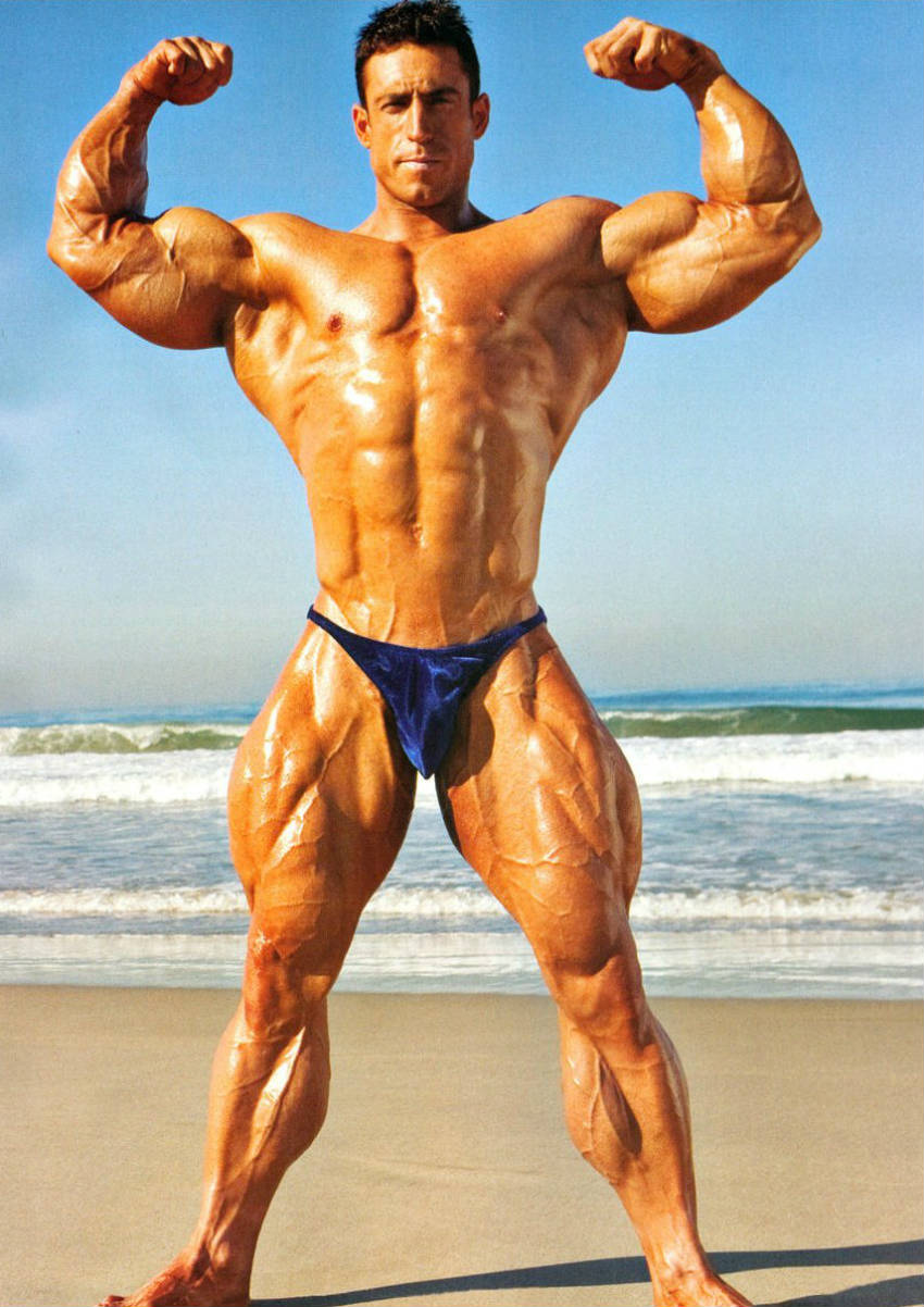 Erik Fankhouser standing on the beach, showing his front profile and tensing his large biceps