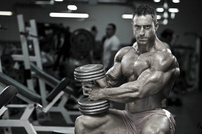 Erik Fankhouser in a black and white photo, sat on the bench in the gym holding a dumbbell and flexing his bicep