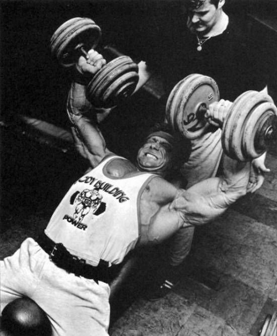 Frank Richards completing a dumbbell bench press with a spotter above him, showing his massive chest and large arms